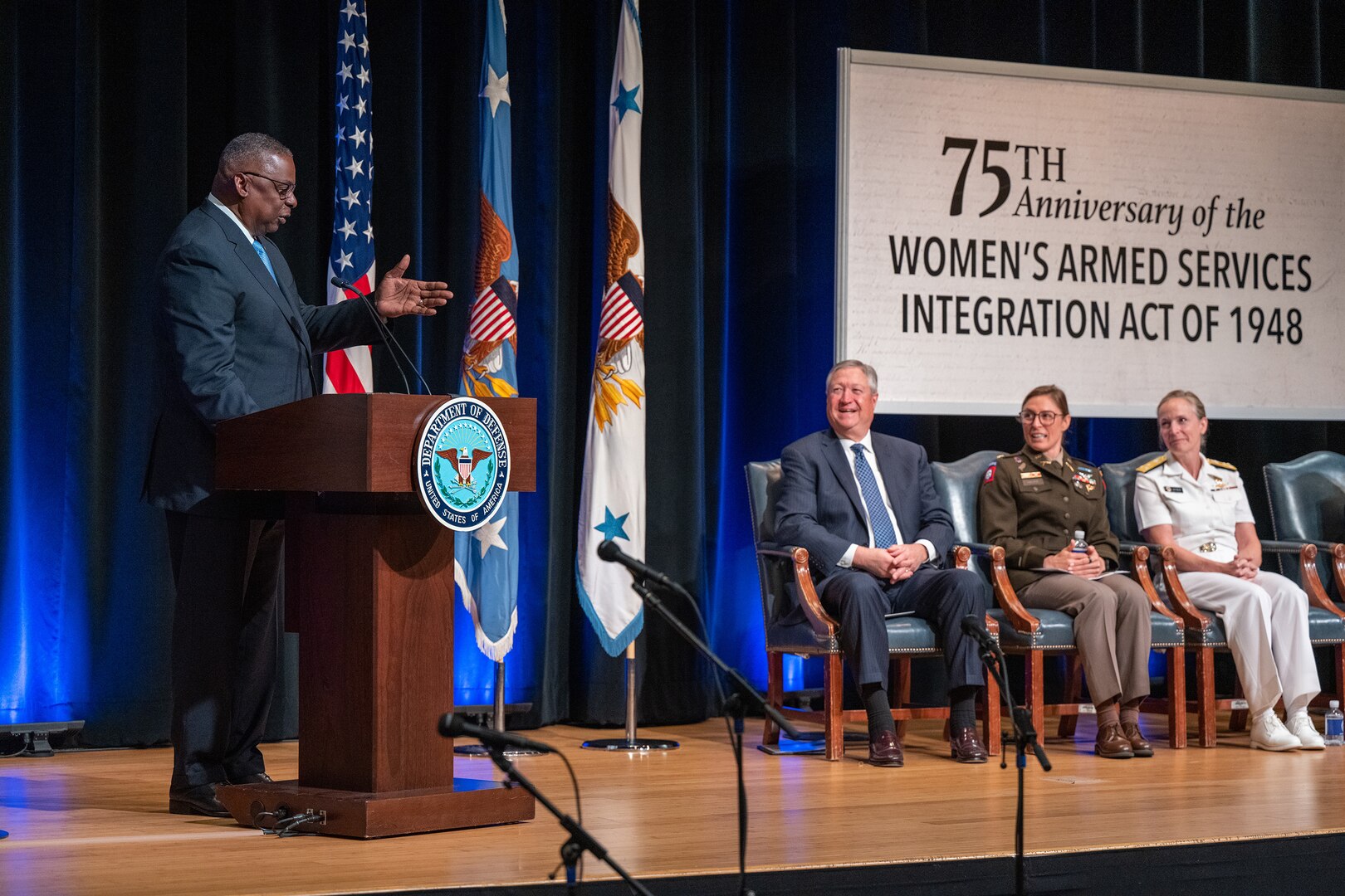 Secretary of Defense Lloyd J. Austin III stands and speaks at a lectern on stage as others seated nearby look on.
