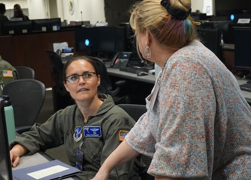 photo of a uniformed U.S. Air National Guard Airman sitting at table filled with computers while a civilian is standing talking to her
