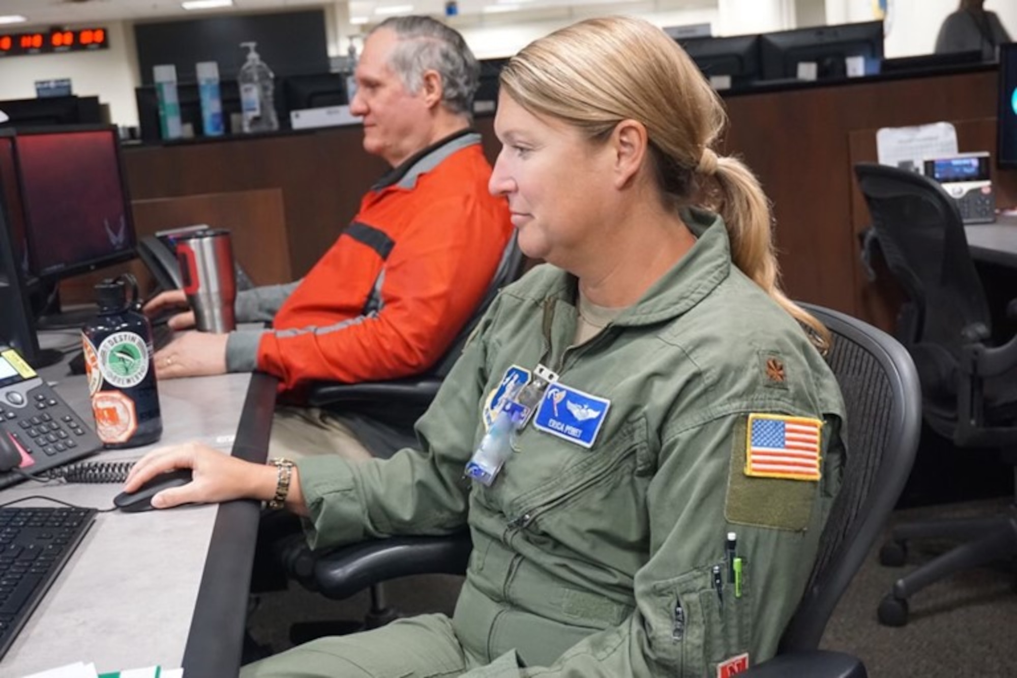 photo of a uniformed U.S. ANG Airman sitting at table filled with computers with a civilian sitting in background working at computer