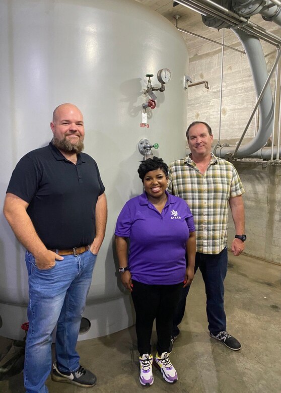 Two men and a woman stand in front of an oil accumulator tank