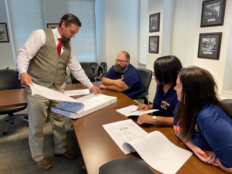 Man stands behind table with document explaining contents to three teacher seated at the table.