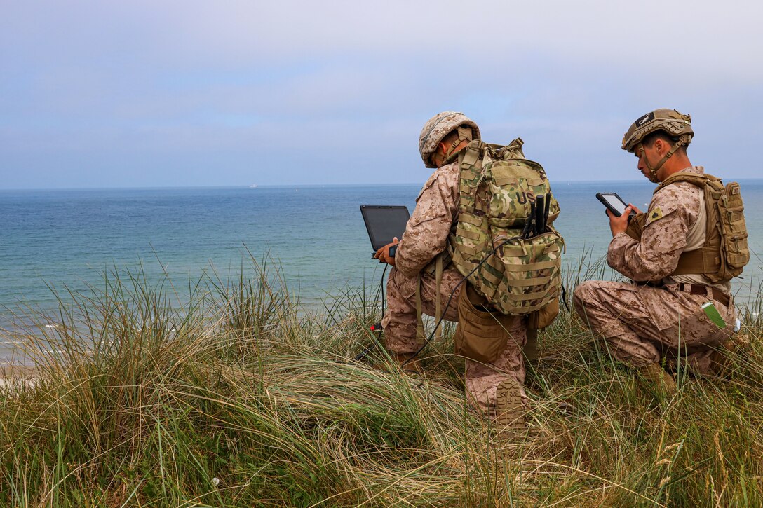 Two Marines take a knee while using digital devices near a shoreline.
