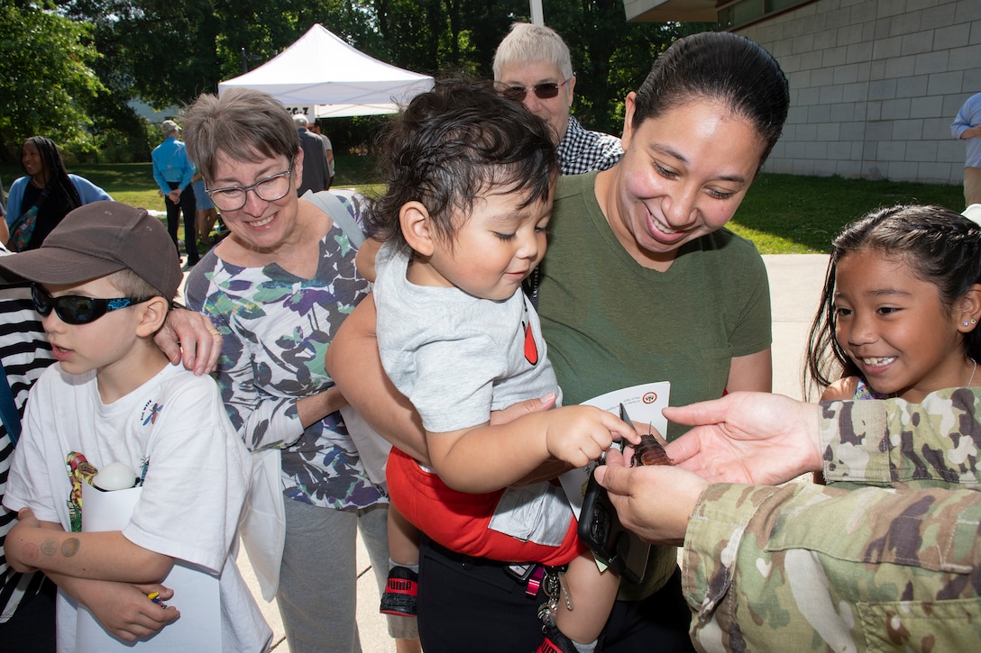 A service member displays an insect for a toddler to touch as others look on, smiling.