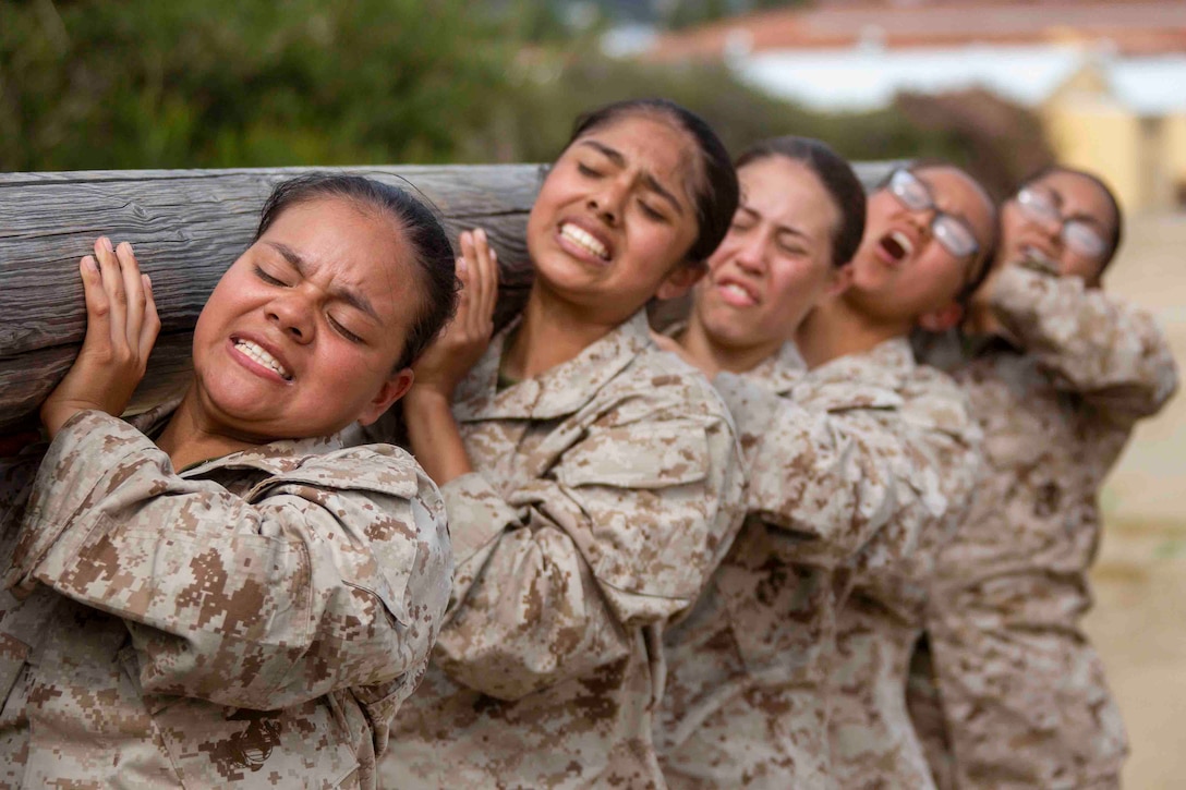 Marine Corps recruits grimace while carrying a log in line on their shoulders.