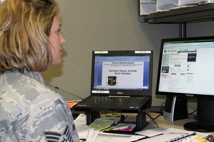A service member sits at a desk with a laptop and a computer screen.