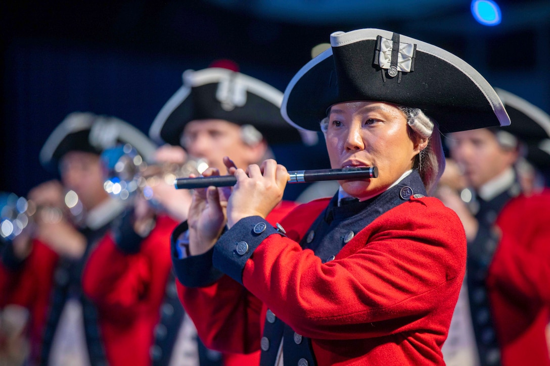 A closeup of a soldier dressed in a ceremonial uniform playing an instrument as fellow soldiers perform in the blurred background.