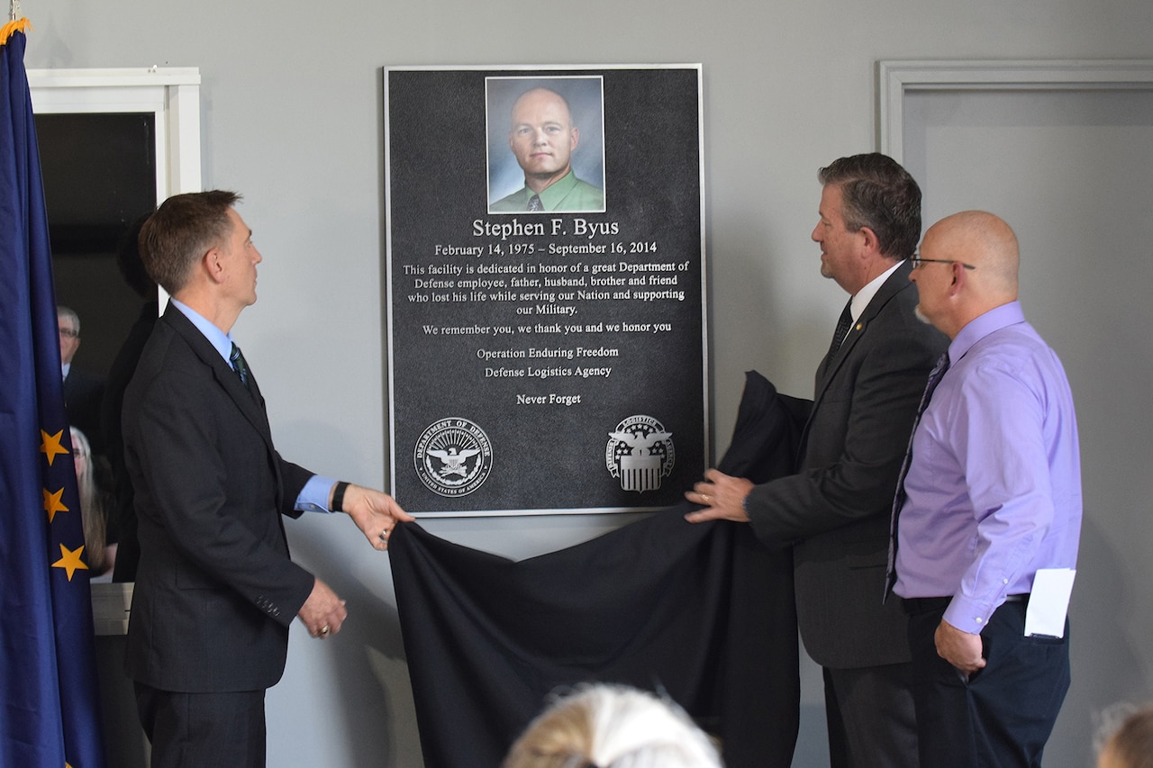 Two people unveil a memorial plaque while another man looks on.