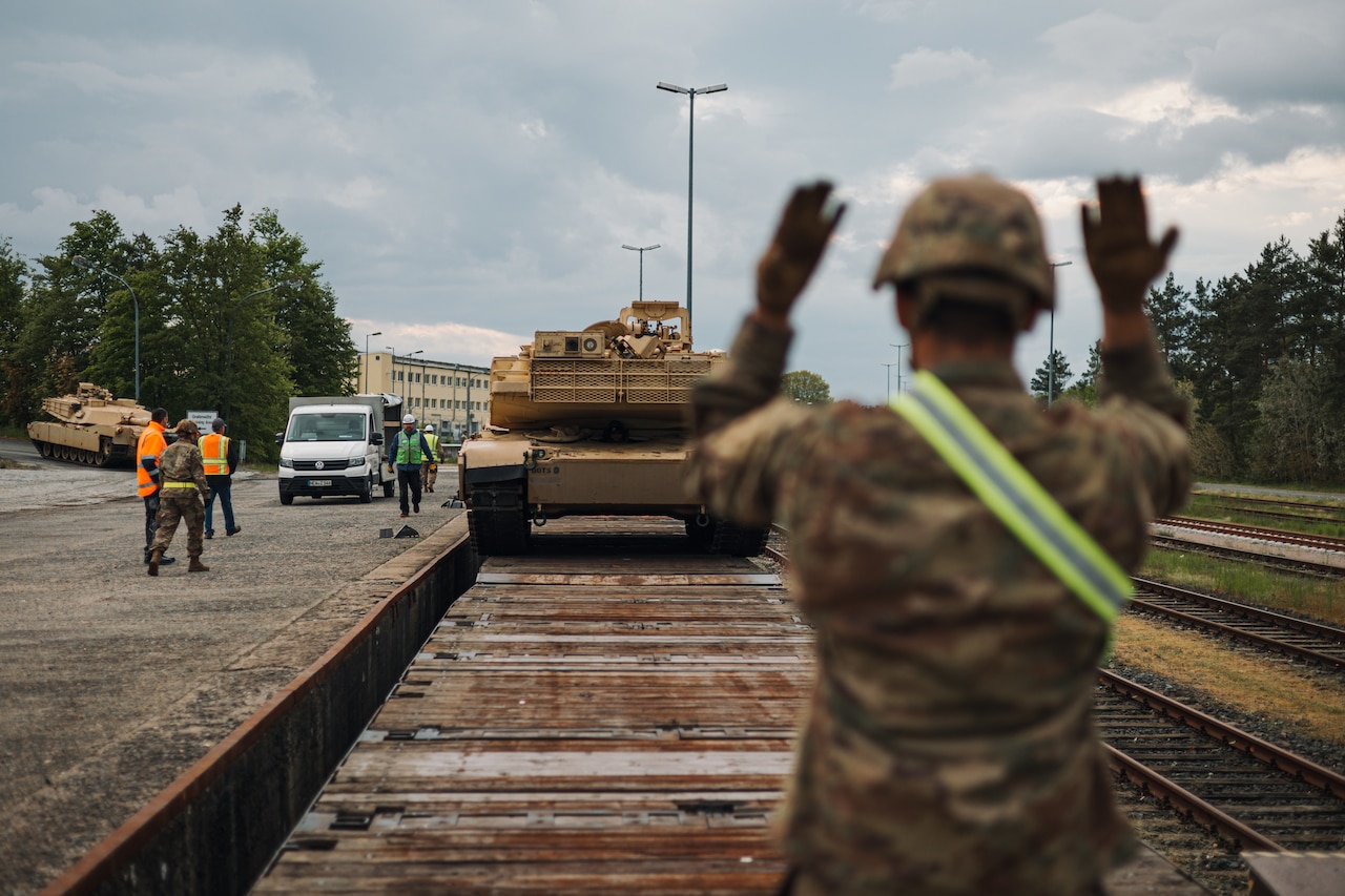 A service member in uniform motions with arm signals toward a tank.