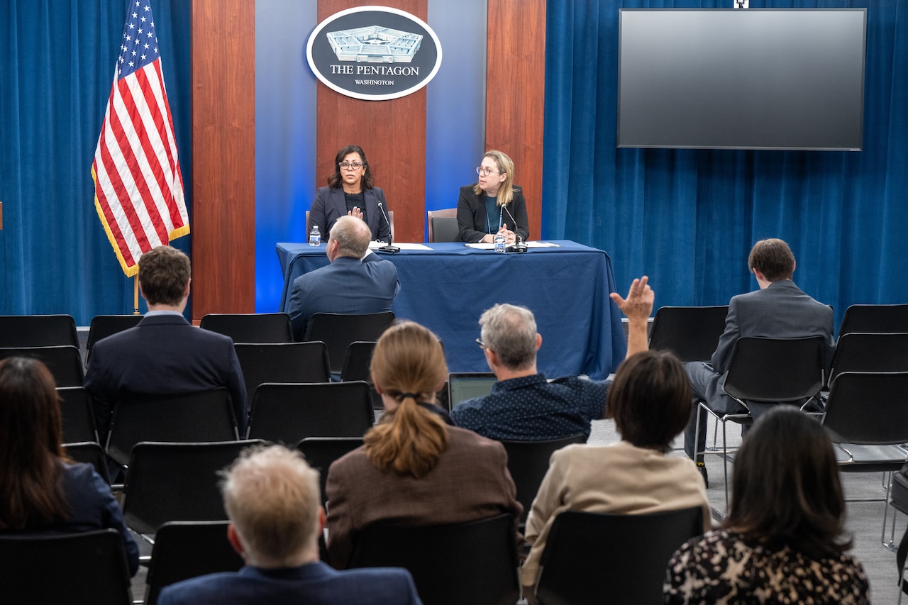 Two women sit at a table. Behind them is an American flag and a sign that says "The Pentagon."  A handful of people sit in chairs in front of them. One raises his hand.