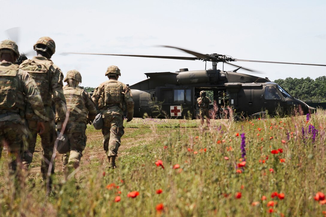 Soldiers walk in a line toward a helicopter with another soldier standing by the helicopter waiting for them.