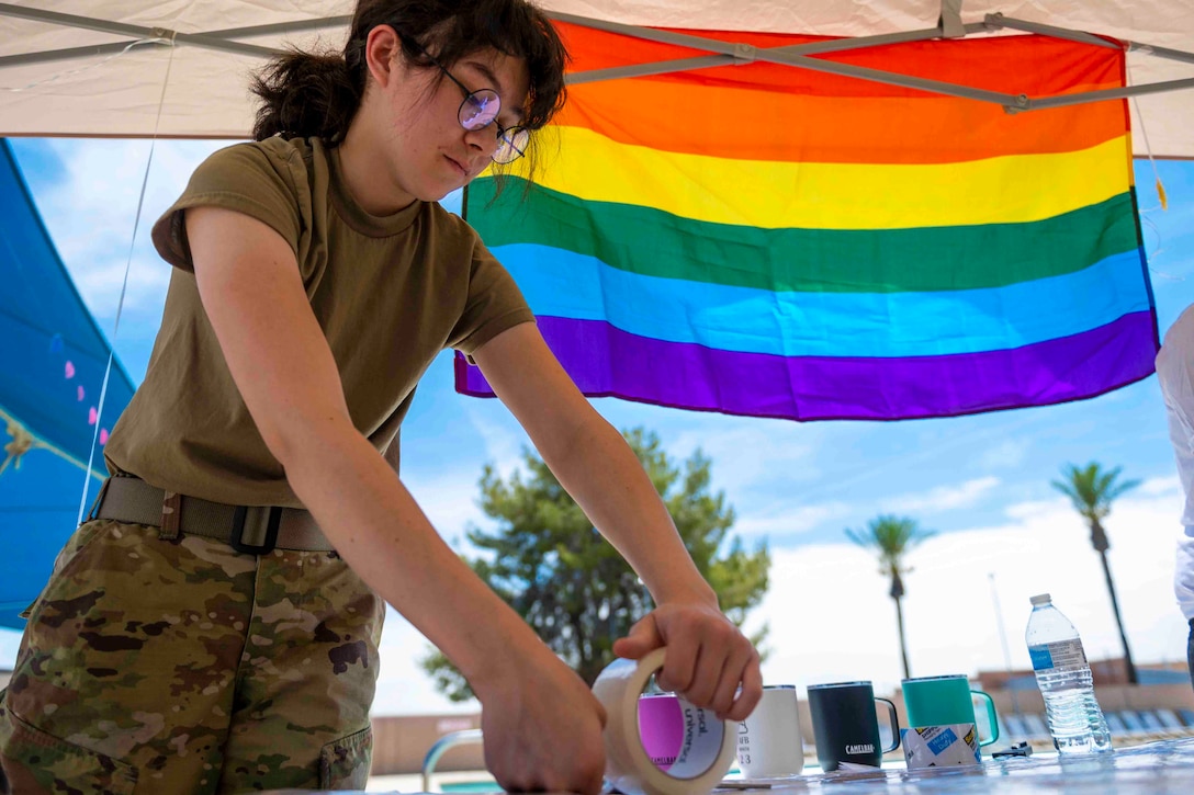 An airman puts tape onto cardboard while standing in front of a rainbow flag.