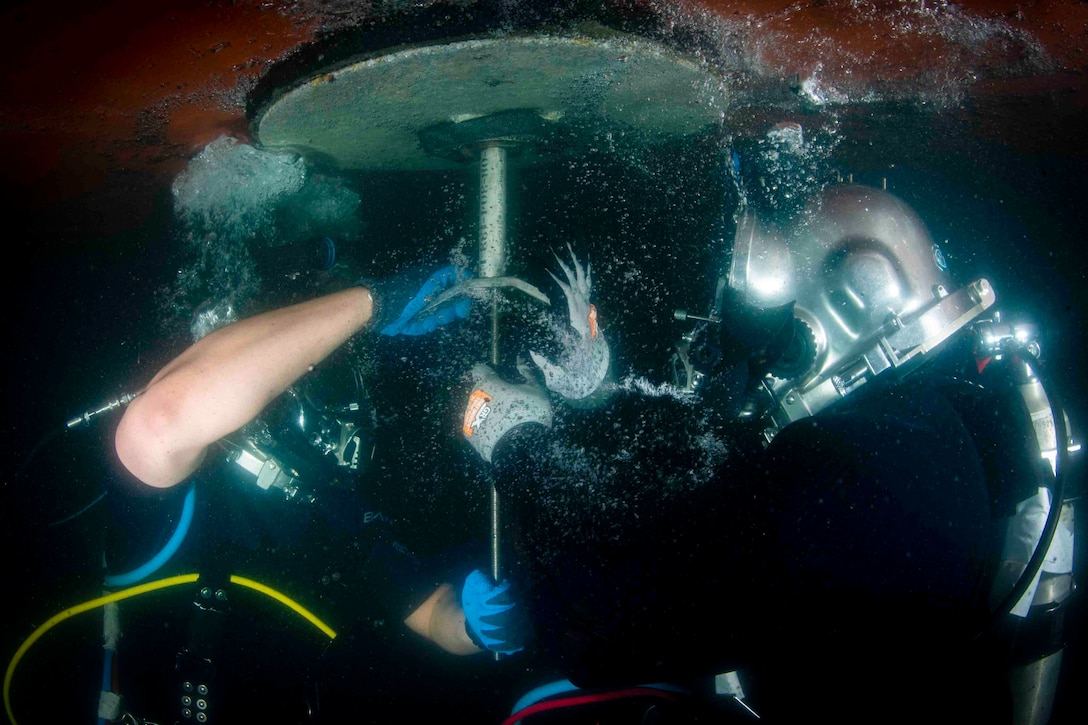 Two sailors in dive gear install a cofferdam on a ship underwater.