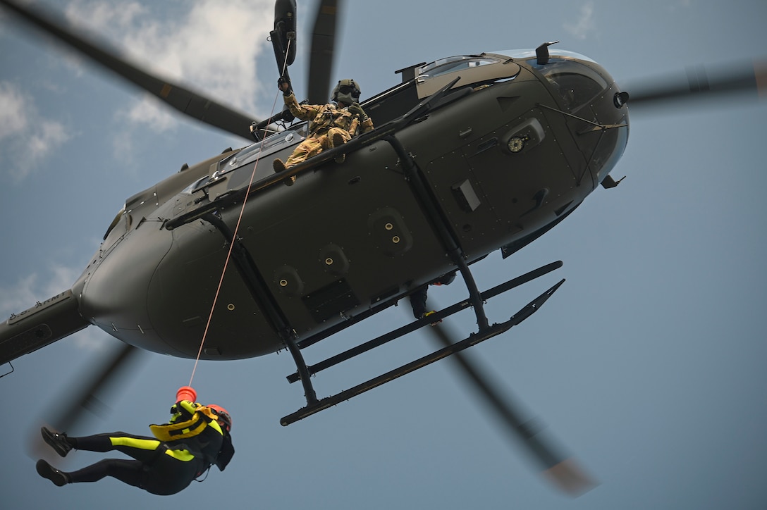 A helicopter flies with a soldier looking out as a person holds onto a hoisting line.