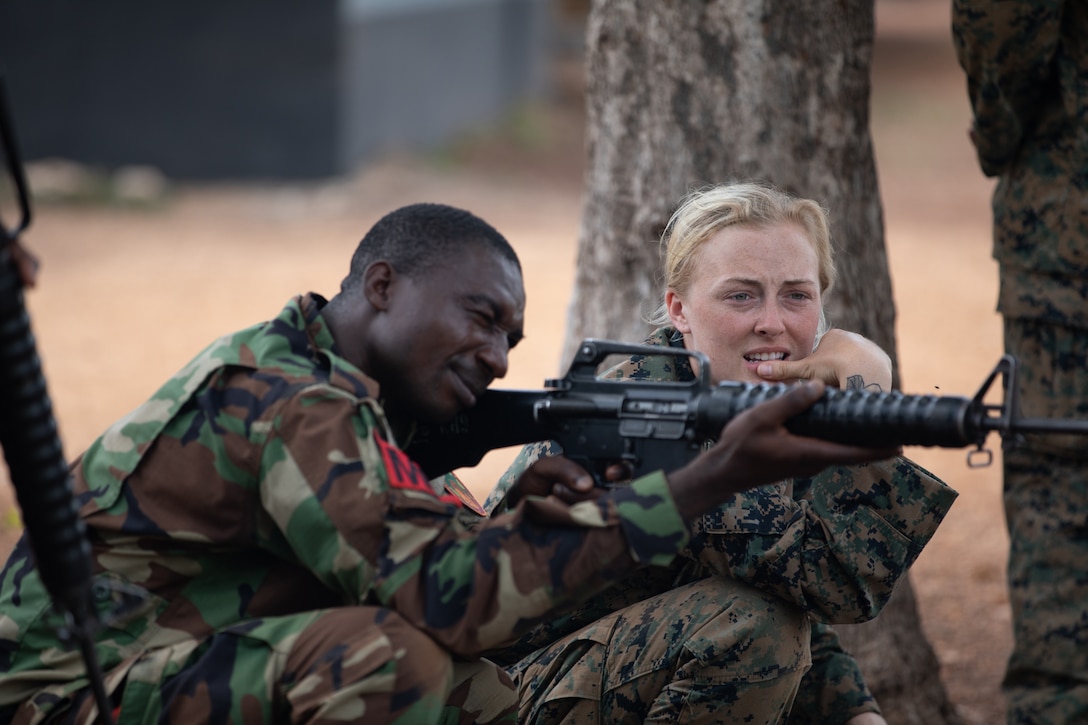 A soldier aims his weapon while a Marine looks on.