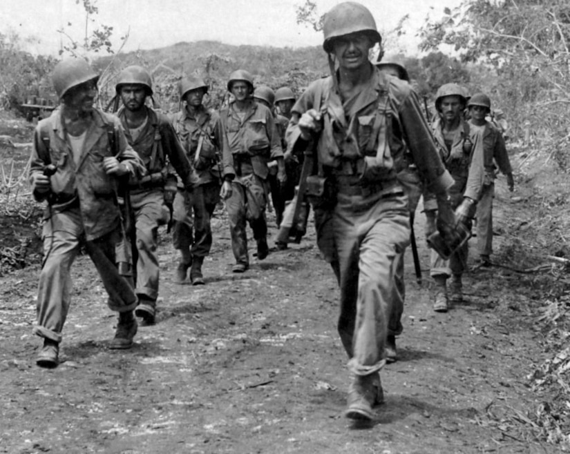 Several men carrying rifles over their shoulders march along a dirt path.