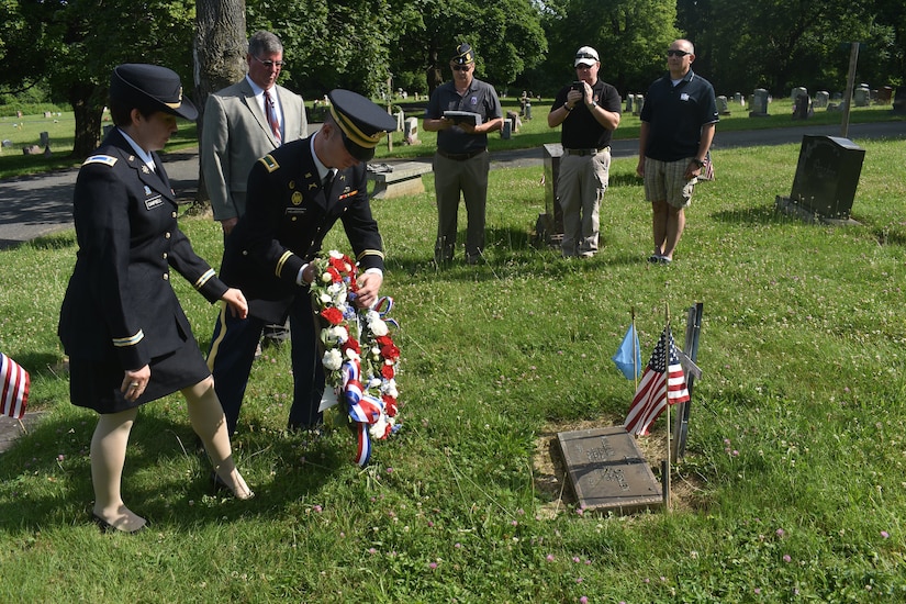 Two people place a wreath in front of a grave as a few others watch in the background.
