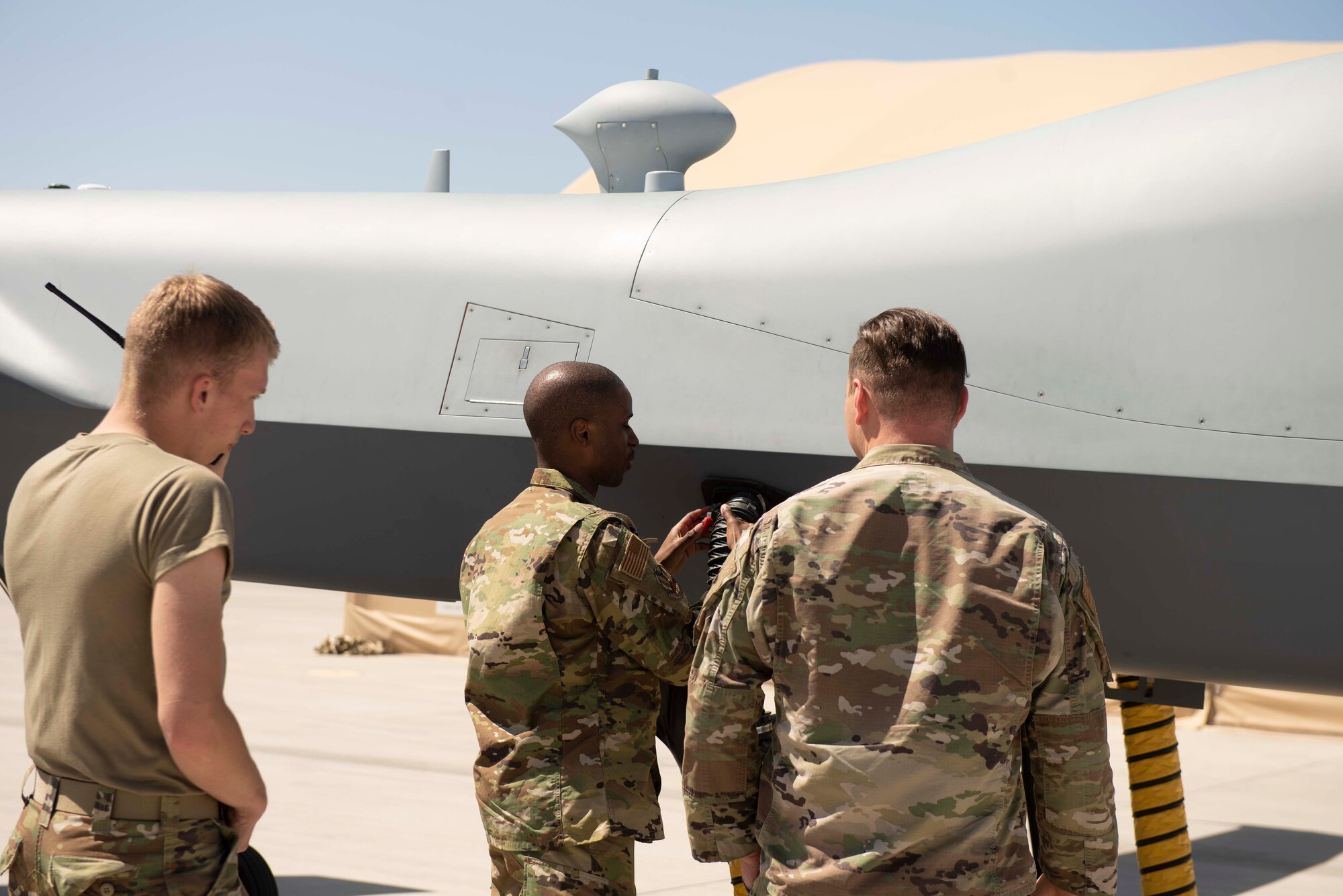 A man in a military uniform operates on a panel on an unmanned aircraft while two other military men observe him.