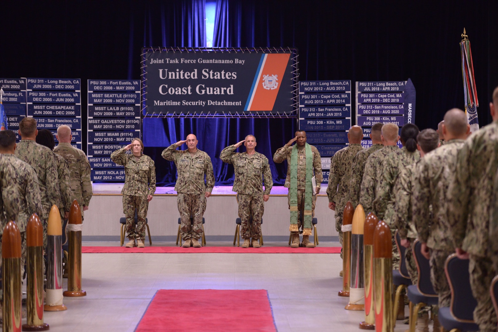 The official party for the U.S. Coast Guard Port Security Unit 305 decommissioning and casing of the colors ceremony, comprised of multiple military branches in the Joint Task Force Guantanamo (JFT-GTMO), stand in front of placards representing all the units that served in the JFT-GTMO mission and salute as the color guard posts the colors at Naval Station Guantanamo Bay, Cuba, June 13, 2023.

PSUs are a Coast Guard deployable specialized force that have served JTF-GTMO as the Maritime Security Detachment for over 21 years. The Virginia-based PSU was the first deployed to Guantanamo Bay in 2002 in support of Operation Enduring Freedom, and this tour completes their fifth unit deployment. (U.S. Coast Guard photo by Petty Officer 1st Class Valerie Higdon)