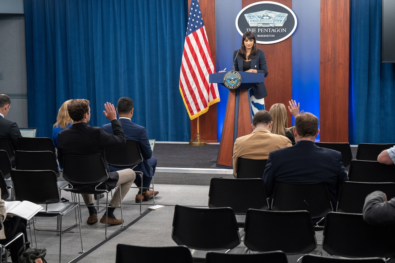 A civilian standing at a lectern addresses a seated audience, some with hands raised.