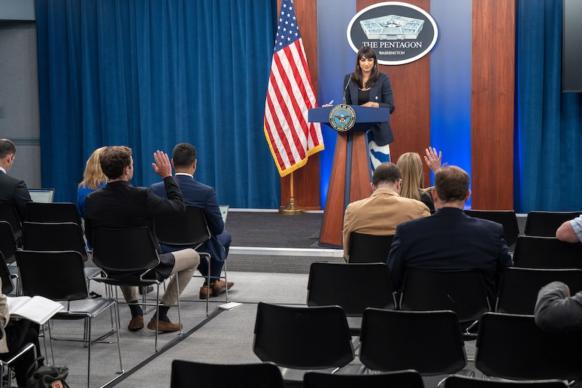 A civilian standing at a lectern addresses a seated audience, some with hands raised.