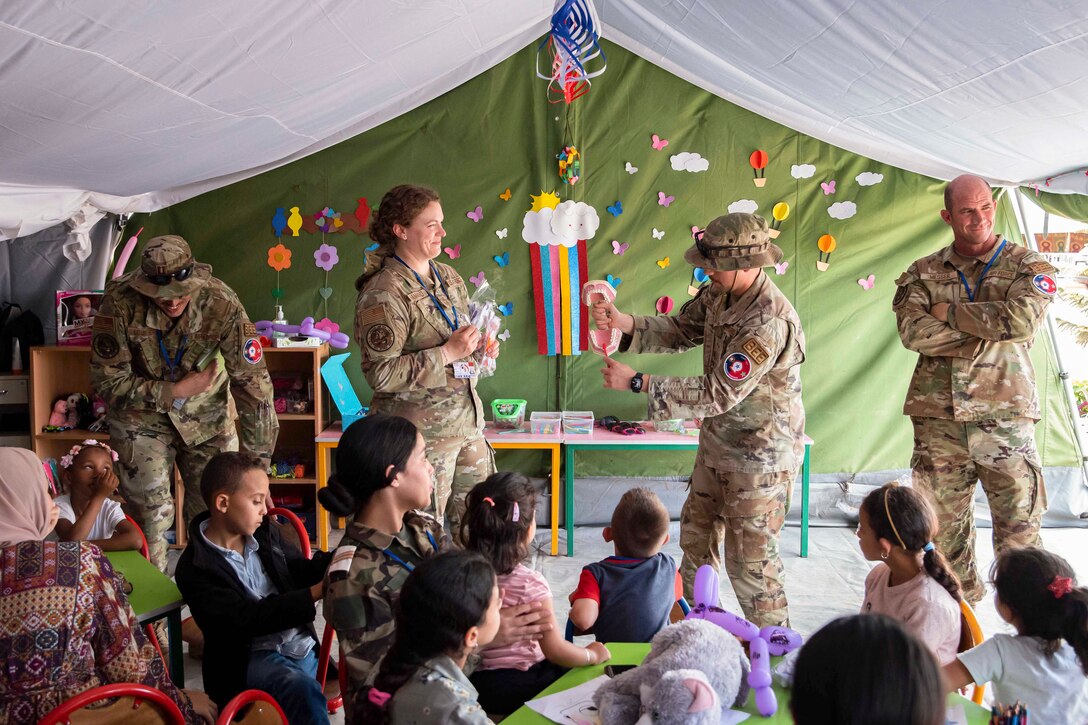 A guardsman standing in a classroom brushes a mock mouth’s teeth as children watch.