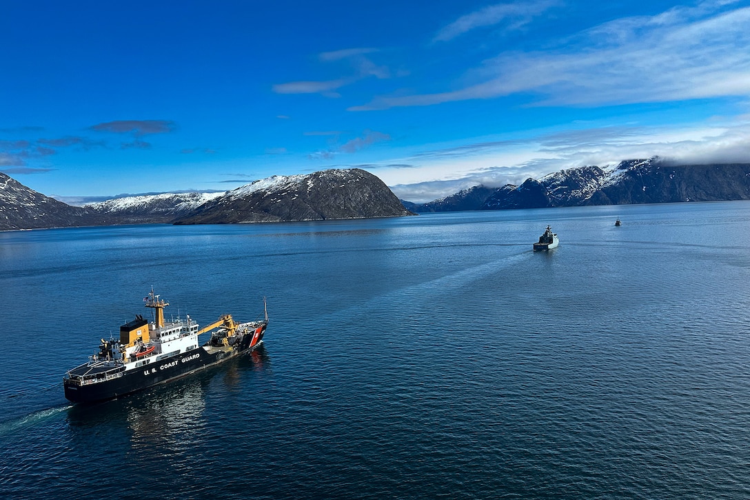 Three military vessels sail in open water with snow-capped mountains in the background.