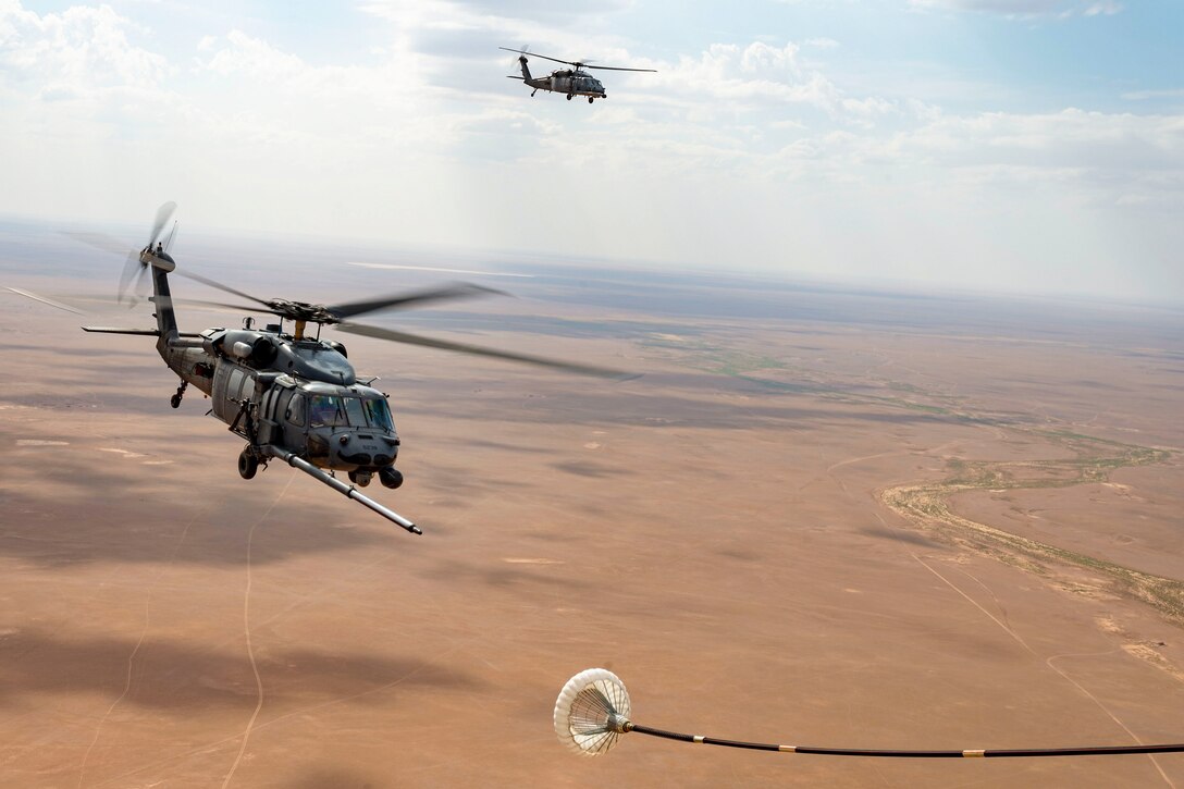 An aircraft's boom extends to a helicopter as another helicopter flies nearby over a desert-like area.