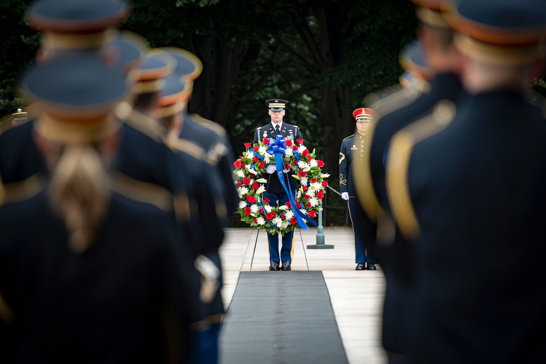 A soldier holding a floral wreath stands in front of fellow soldiers in ceremonial uniform standing in formation during a ceremony.