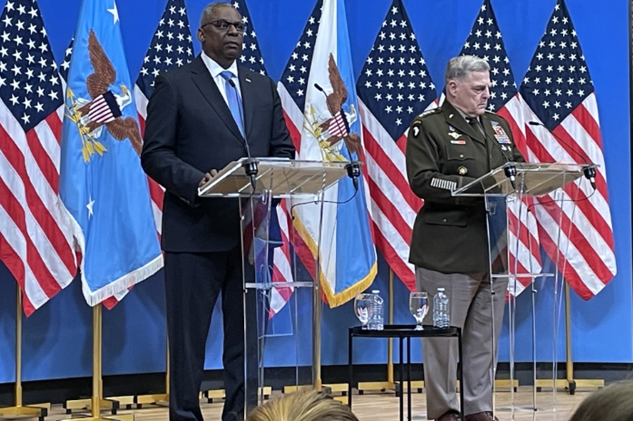 Two men stand behind lecterns with U.S. flags in the background.
