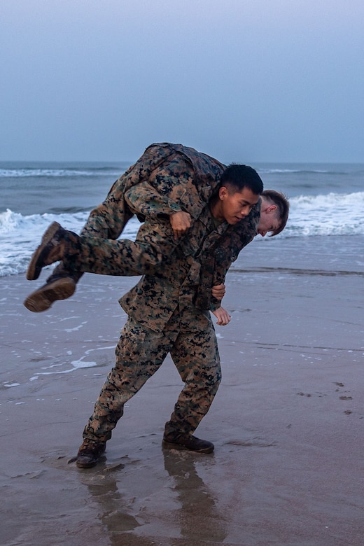 A Marine carries a fellow Marine while running on the beach.
