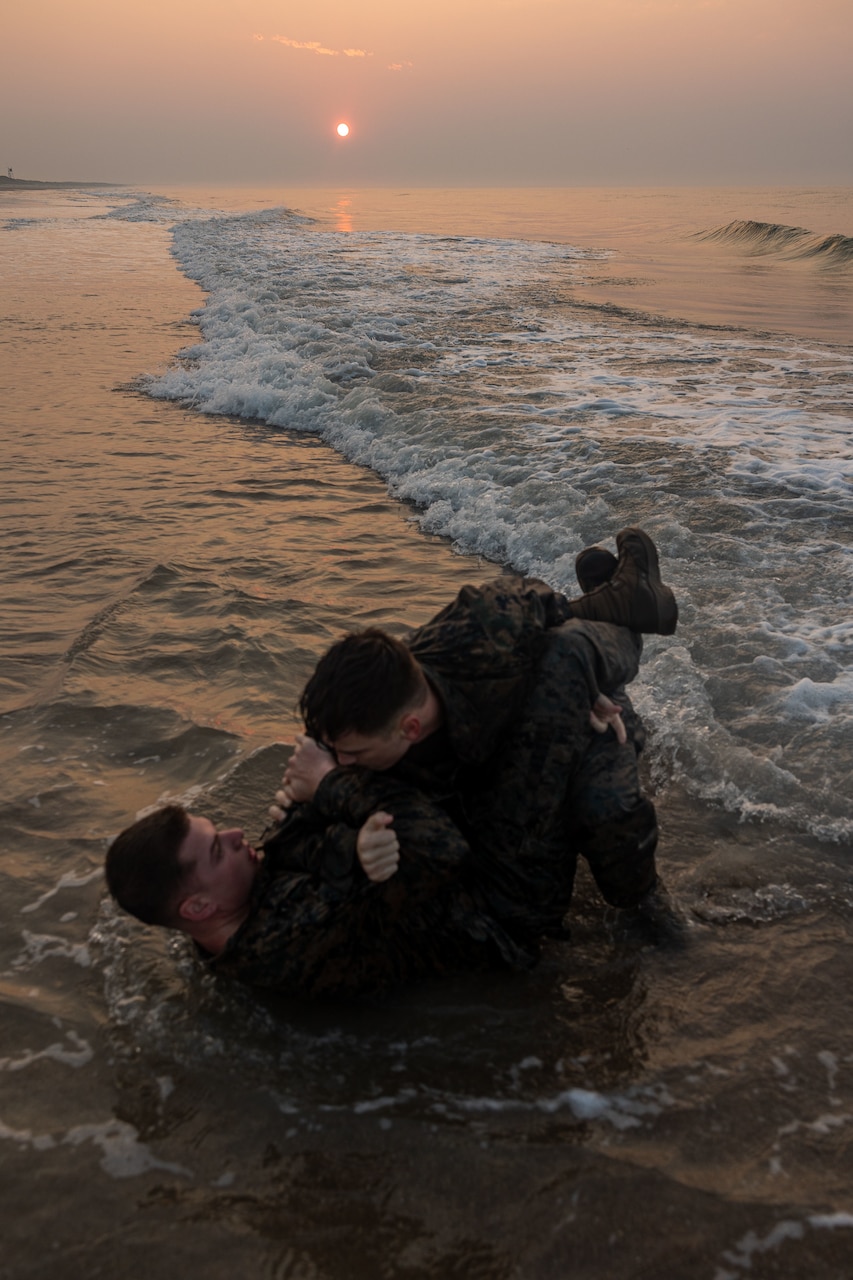 Two Marines grapple on the beach under a sunlit sky.
