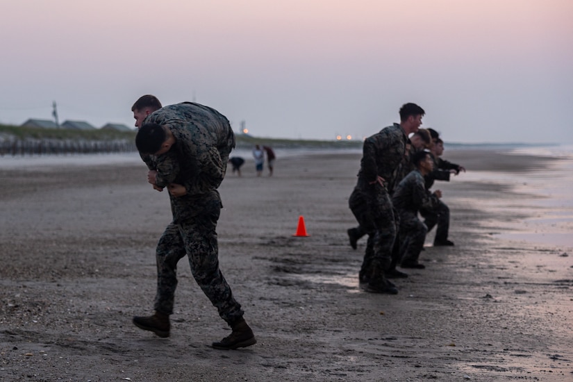 Marines squat in the sand as a fellow Marine carries another.