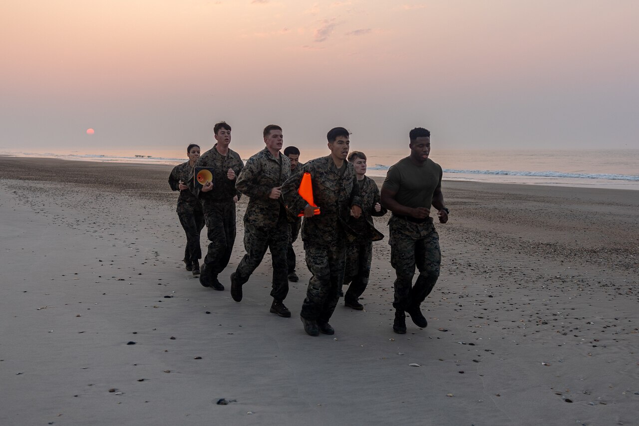 Marines run on the beach under a sunlit sky.