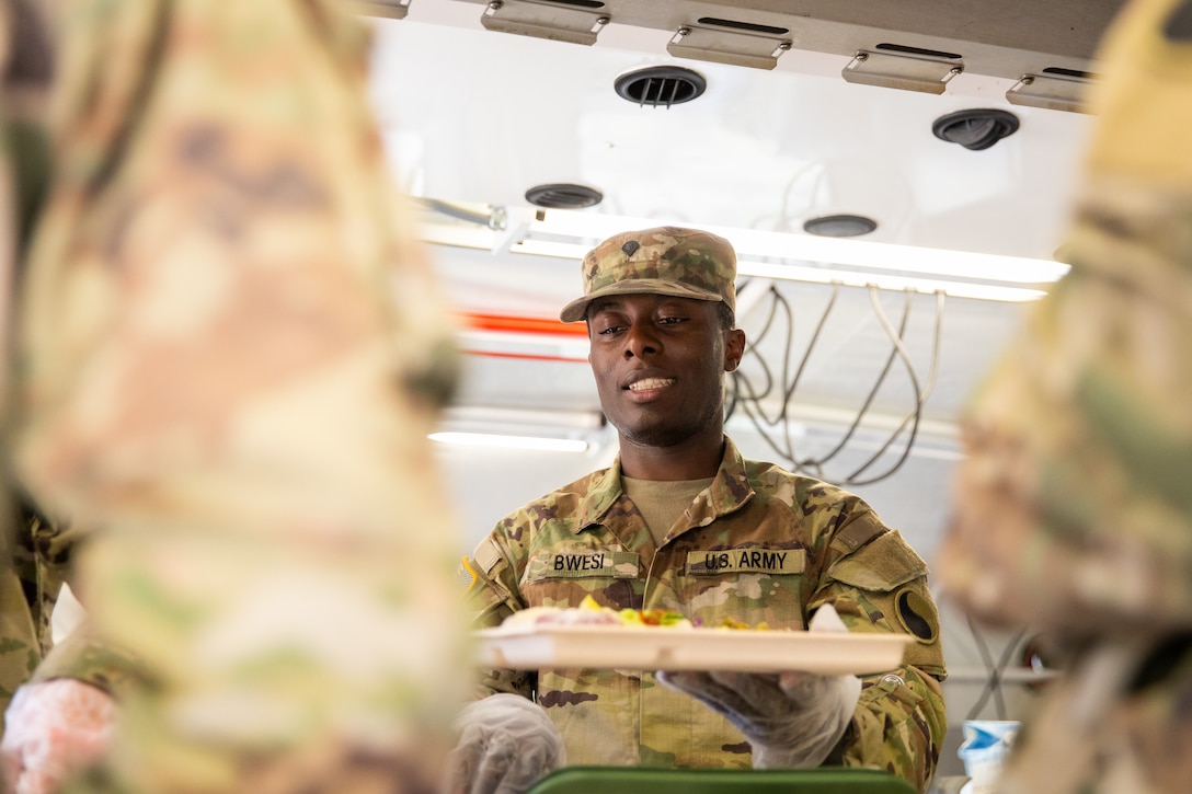 U.S. Army Spc. Adam Bwesi of India Company, 429th Brigade Support Battalion, 75th Troop Command, serves food to a Soldiers in a containerized kitchen during the Regional Philip A. Connelly Field Feeding Competition at the Harlold L. Disney Training Center in Artemus, Kentucky on June 14, 2023. Bwesi and the rest of India Company are competing against nine other states and territories National Guard field feeding units in the southeast United States for a chance to compete nationally against the other regions of the National Guard. (U.S. Army National Guard photo by Andy Dickson)