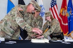 Three military members in camouflaged uniforms cut a white cake on a table.