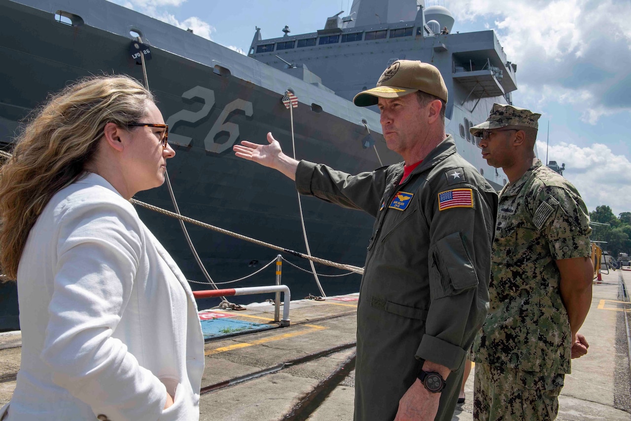 A man in military uniform points to a ship while another man in uniform stands behind him. A woman looks on.