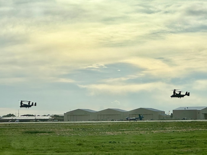 Aircraft sits center on the flight line against a background of aircraft hangars while helicopters fly in the air on each side of the canvas