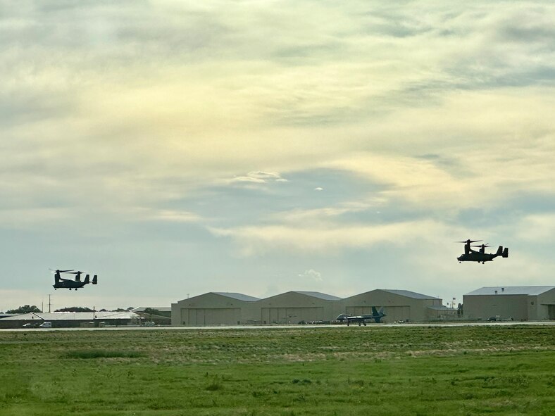 Aircraft sits center on the flight line against a background of aircraft hangars while helicopters fly in the air on each side of the canvas