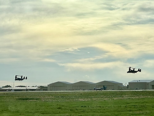 Aircraft sits center on the flight line against a background of aircraft hangars while helicopters fly in the air on each side of the canvas
