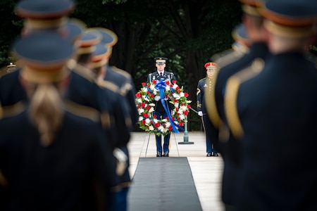 A soldier holding a floral wreath stands in front of fellow soldiers in ceremonial uniform standing in formation during a ceremony.