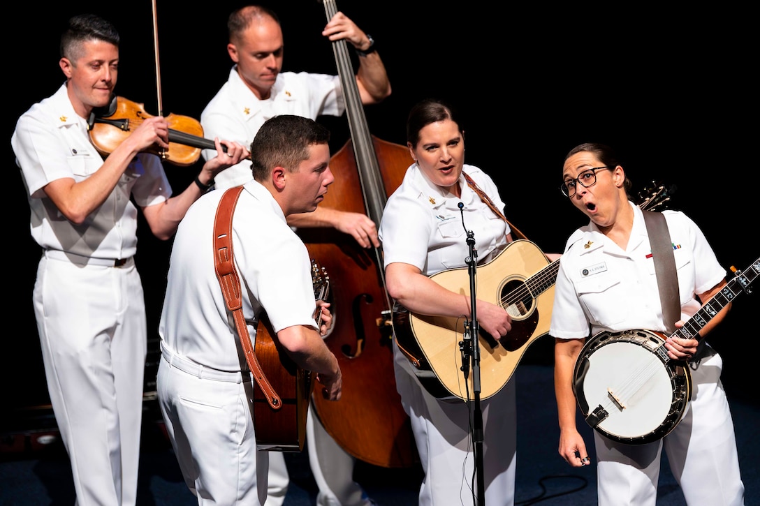 Five sailors play instruments on a stage; some sing into a microphone.