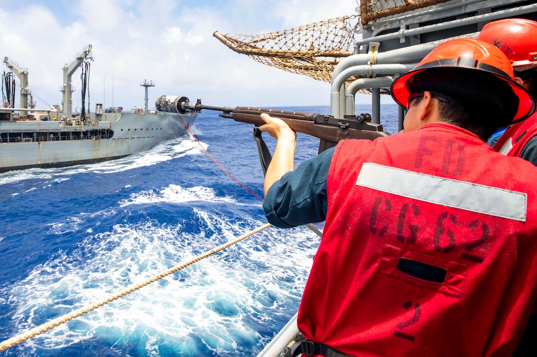 A sailor standing on a ship prepares to fire a line at another ship sailing nearby.