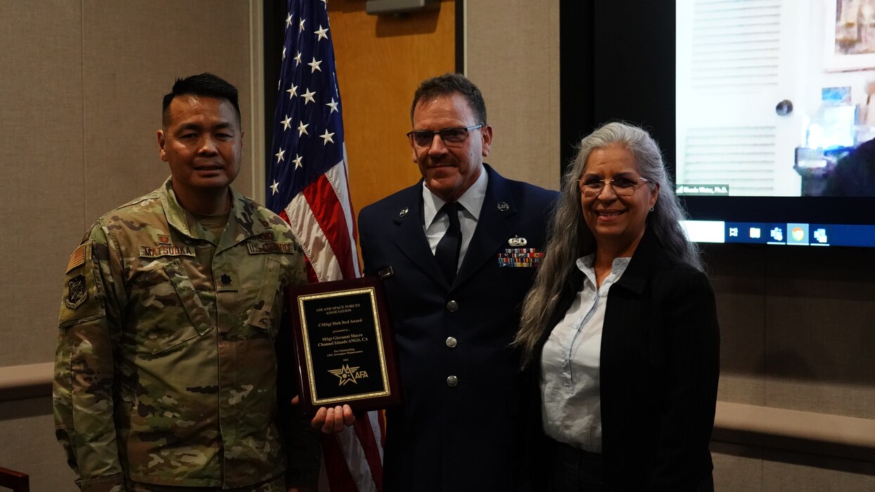 A photograph of Senior Master Sgt. Giovanni Macro posing for a photo with his award inside a room next to his wife and his supervisor.