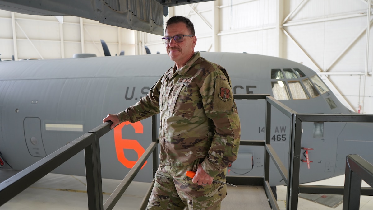 A photograph of Senior Master Sgt. Giovanni Macro posing for a photo on top of an aircraft lift inside a large aircraft hangar with a C-130J Super Hercules behind him.