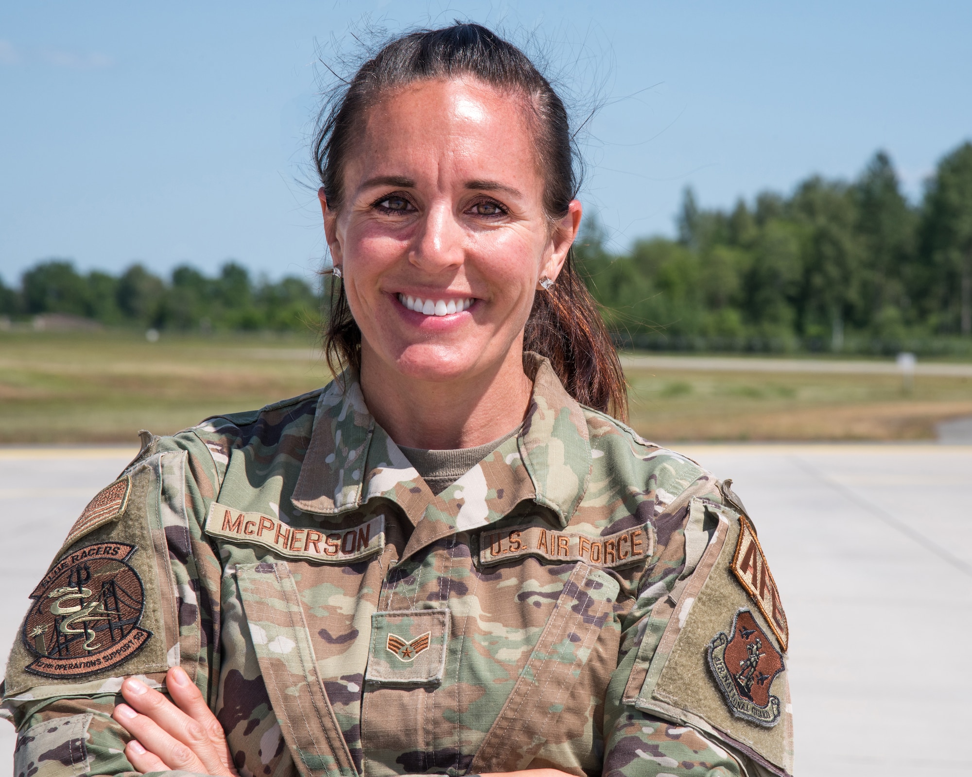 Female Airman wearing fatigues poses with crossed arms in front on a concrete flight line, forest trees in the background.