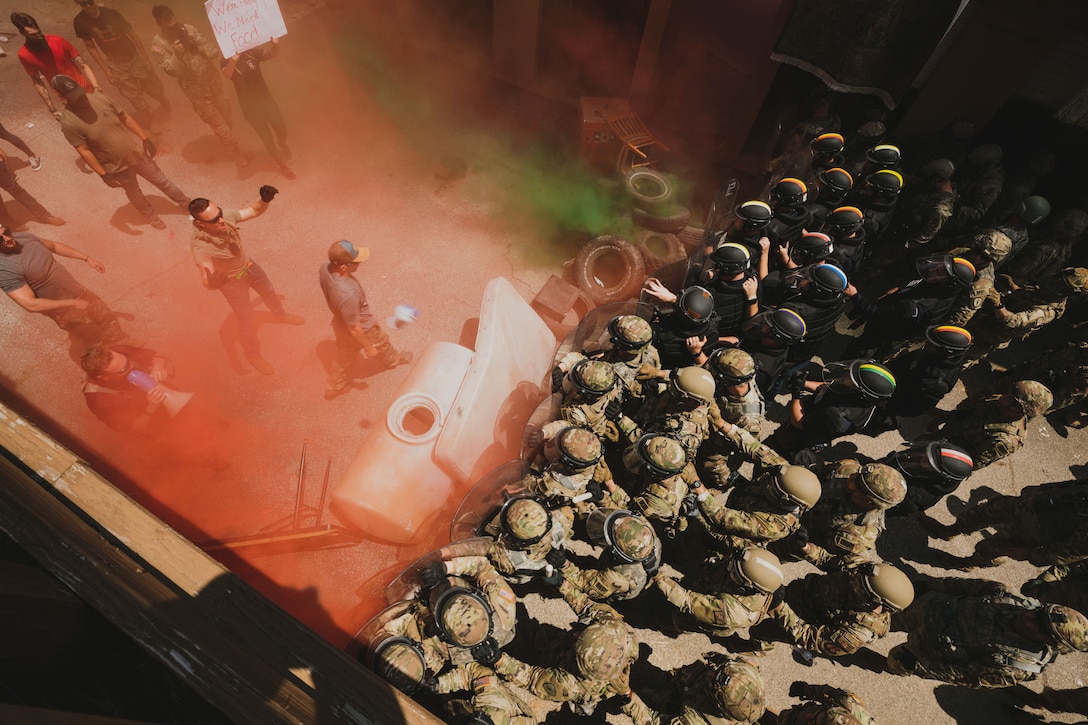 A large group of soldiers and police crowd together and some hold shields as people protest in a smoky area.