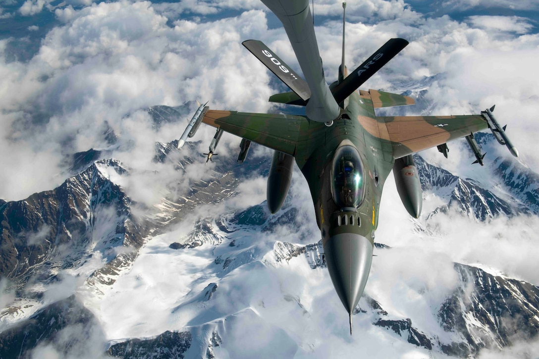 A jet prepares to receive fuel midair above snow-covered mountains.