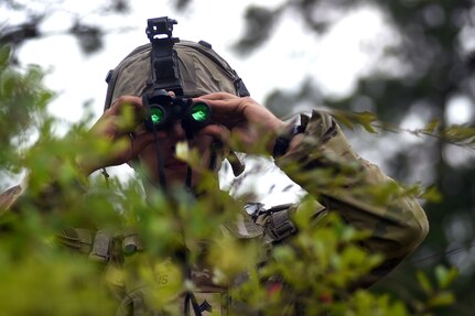 Spc. Grant Simmons, a combat engineer with the 1123rd  Engineer Company (Sapper), Kentucky Army National Guard, uses binoculars to search for enemy movement after his unit was attacked during a training rotation at the Joint Readiness Training Center at Fort Johnson, Louisiana, June 5, 2023. The rotation allowed Soldiers to build cohesiveness and readiness as they trained on large-scale combat operations.