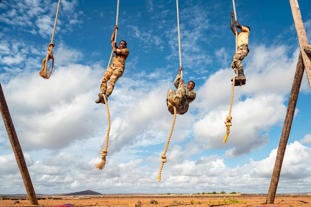 A U.S. soldier and two Djiboutian soldiers climb ropes attached to a wooden obstacle in a desert-like area.
