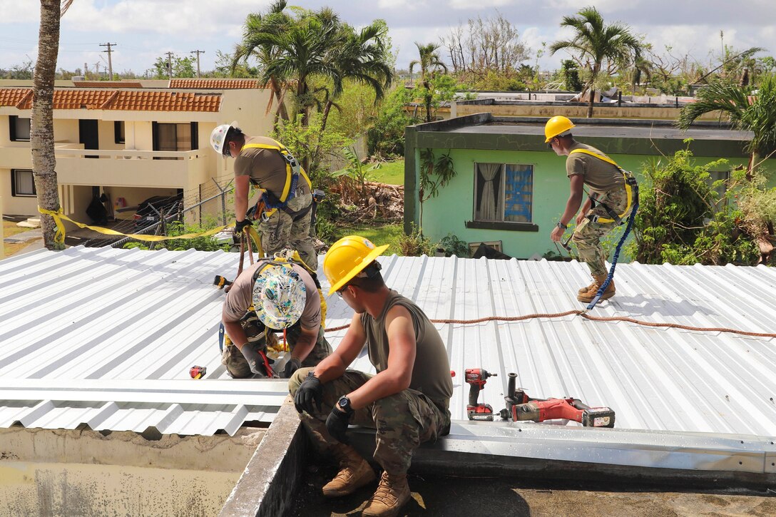 Three soldiers work on the roof of a home as a fellow soldier sits and watches.